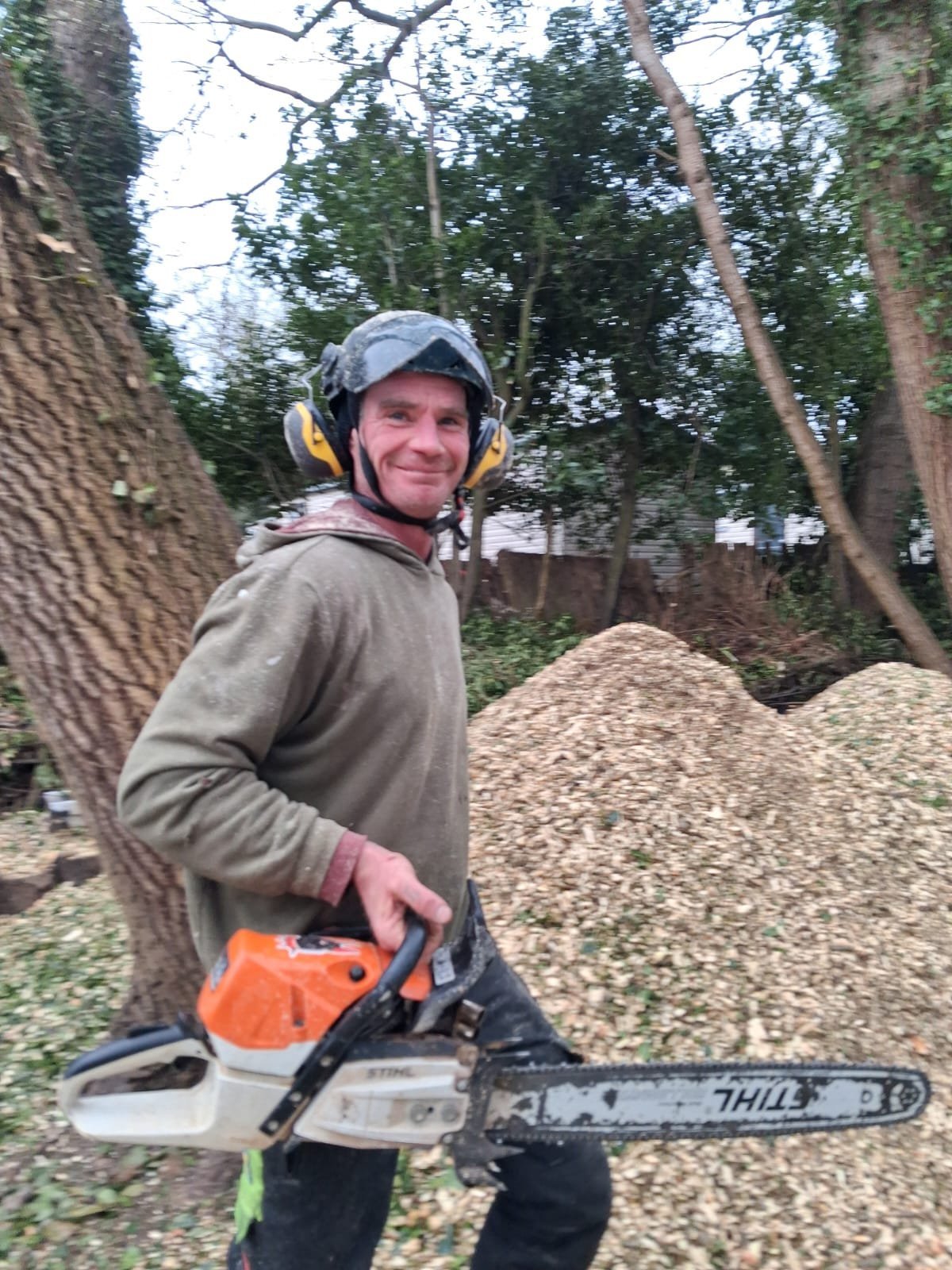 Man holding a chainsaw outdoors wearing safety gear and cap near trees and wood pile